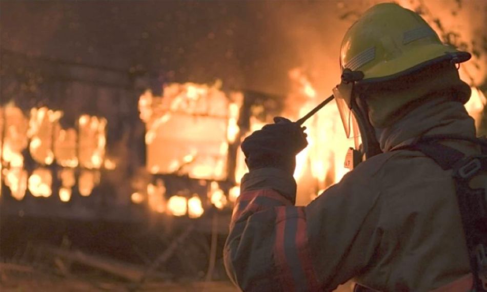 A firefighter using a Tait radio in front of a burning building during an emergency response.